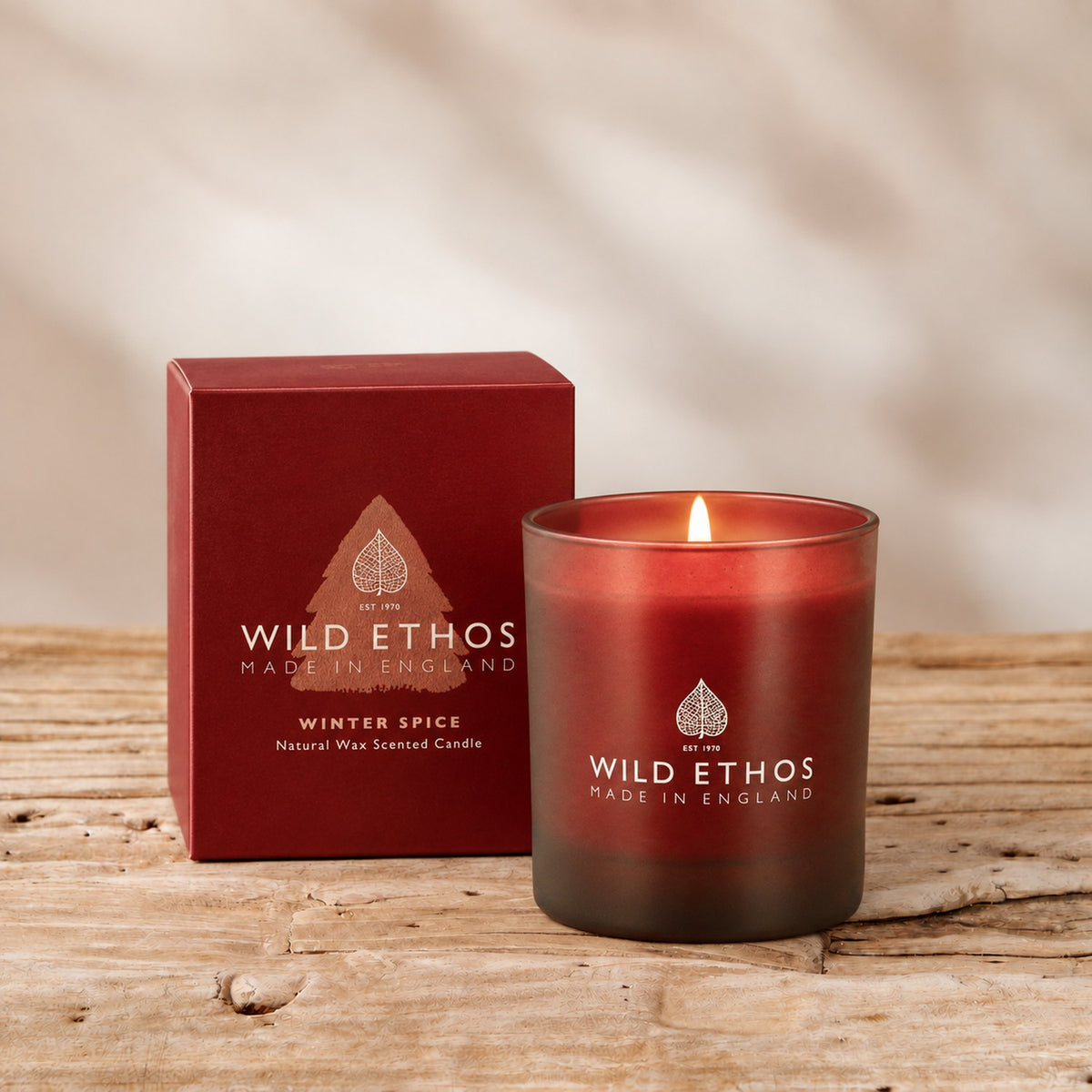 Wild Ethos Winter Spice scented candle burning in a red frosted glass jar beside its red gift box on rustic wood